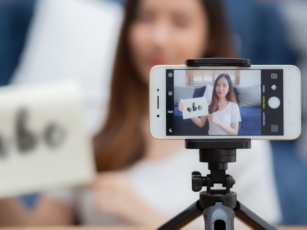 Woman holding up a piece of paper while teaching online. She's blurred in the background with her cell phone on a small tripod showing her more clearly in the foreground.