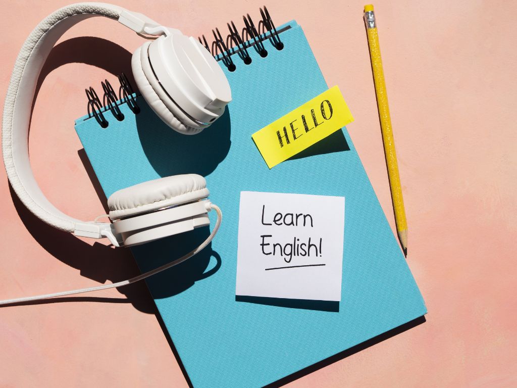 White headphones sitting on top of a teal notepad. Two sticky notes on the pad say Hello and Learn English.