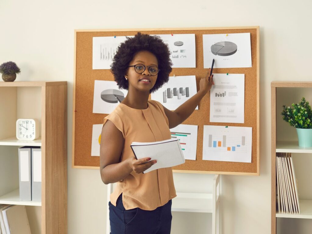 Black female teacher with glasses pointing at printouts on a bulletin board while holding an open notebook in her other hand, demonstrating her transferable skills for teachers.