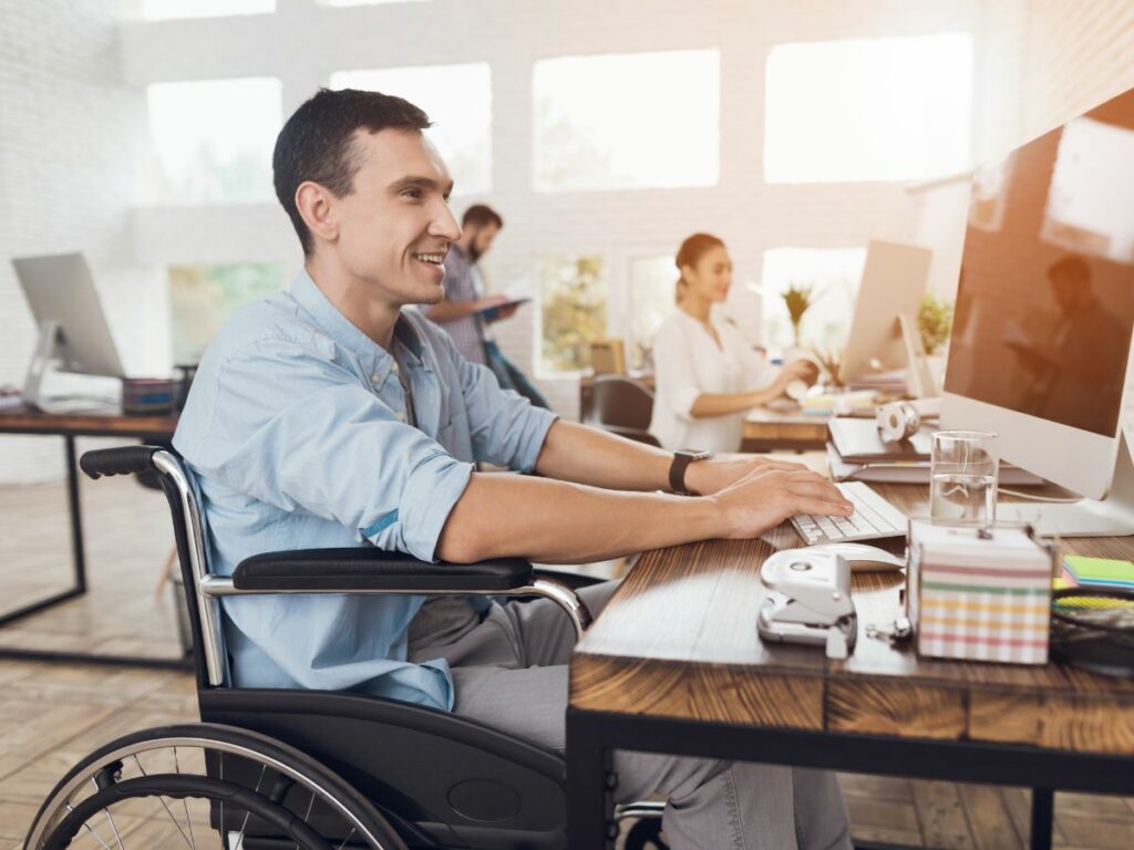 Man in a wheelchair typing at a desktop computer in an open-air office environment.