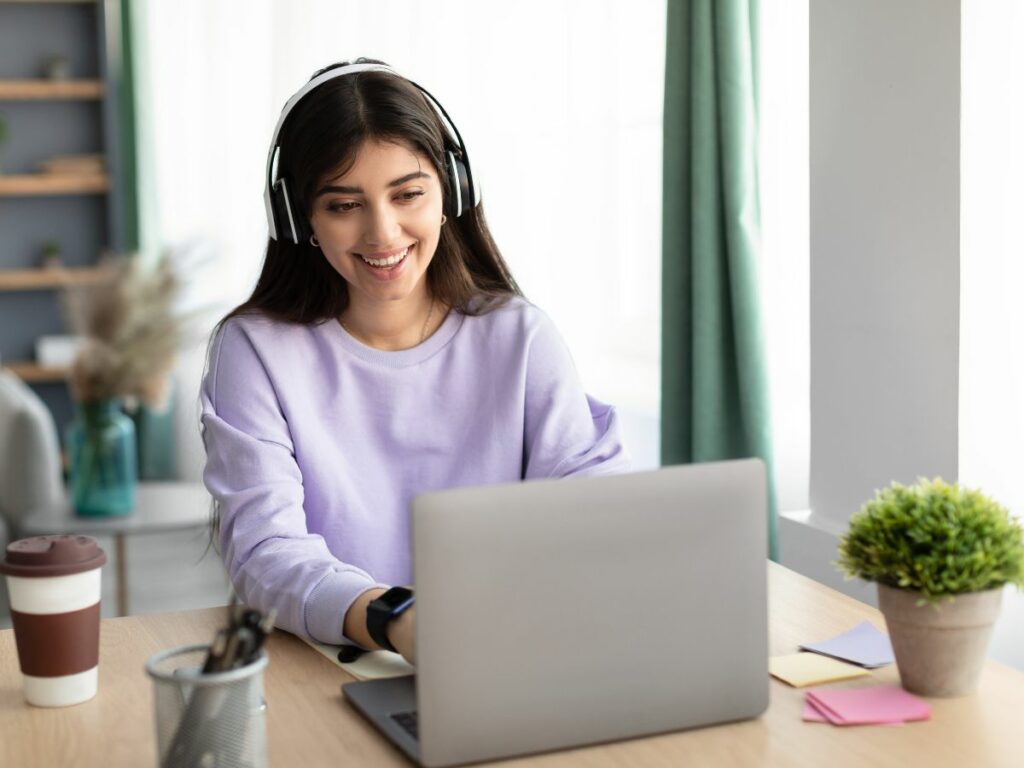 Woman typing on a laptop while wearing headphones and sitting at a desk in a well-lit home office. 