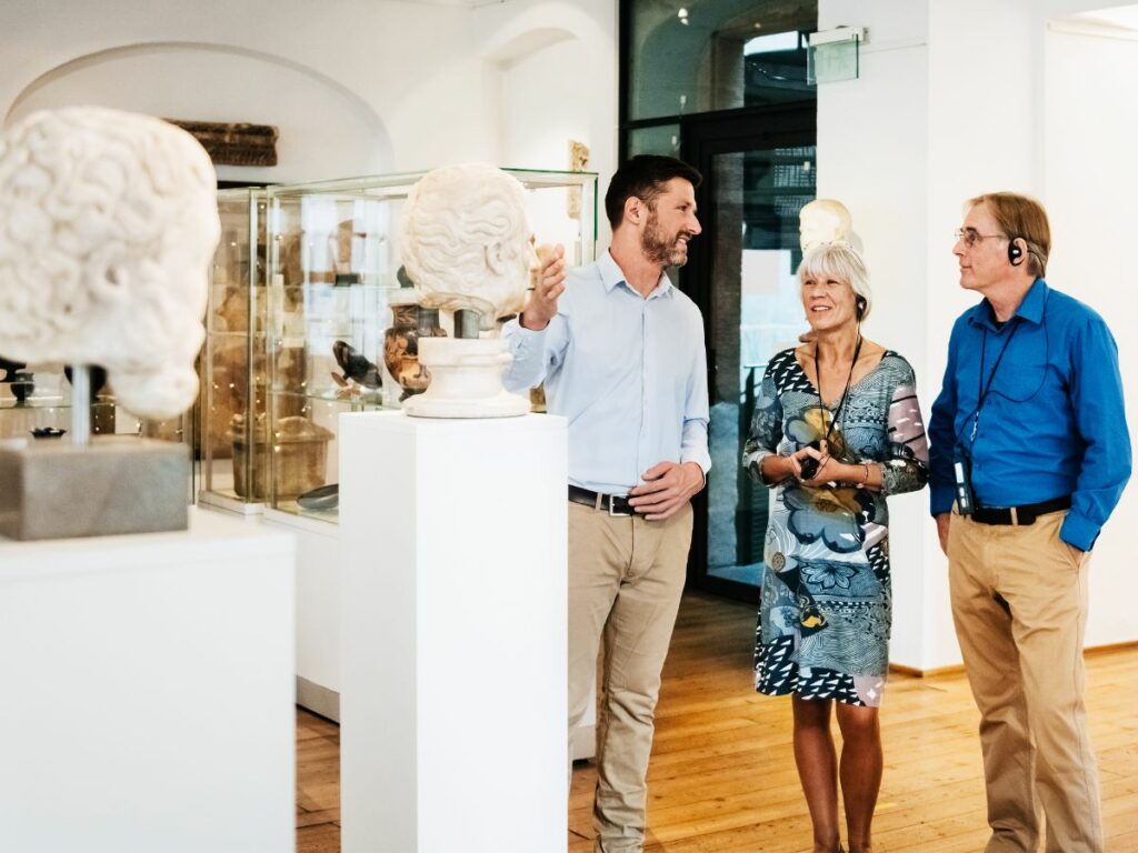 Man talking to two people at a museum while gesturing at a marble bust on display.