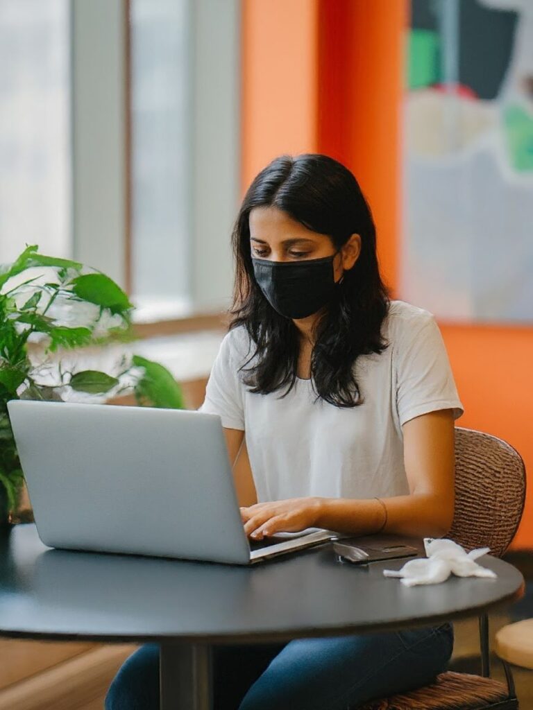 A woman wearing a face mask while working on her laptop in a coworking space, showing how the history of coworking was forever changed by the COVID-19 pandemic.